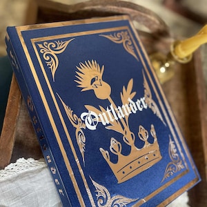 Può includere: Un libro con copertina rigida blu navy e dettagli dorati. Il titolo "Outlander" è scritto in bianco, con un disegno di cardo e corona dorati. I bordi della copertina sono decorati con filigrane dorate.