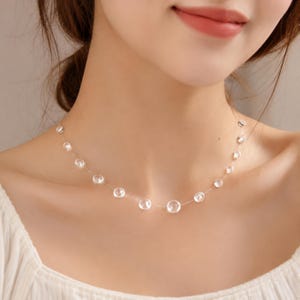 May include: A necklace featuring a series of clear, spherical beads spaced along a thin, transparent nylon cord. The beads vary slightly in size, with the largest bead positioned at the center, creating a floating effect around the neck.