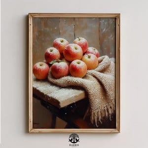 May include: A framed still life painting of red apples dusted with white powder, arranged on a wooden surface with a beige scarf. The apples are the focal point, with a warm, inviting color palette.