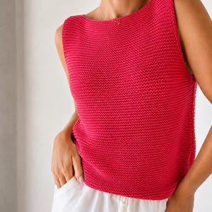 Könnte beinhalten: Ein ärmelloses, leuchtend fuchsiafarbenes Strickoberteil mit texturiertem Kraus-rechts-Muster, weitem U-Boot-Ausschnitt und geradem Saum. Das Kleidungsstück wird mit einer weißen Hose kombiniert, die einen Kordelzug und sichtbare Seitentaschen hat.
