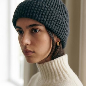 Könnte beinhalten: Eine dunkelgraue Rippstrick-Beanie. Der Hut wird mit einem cremefarbenen Rollkragenpullover getragen. Die Person trägt goldene Creolen. Die Beanie ist ein klassisches Winteraccessoire.