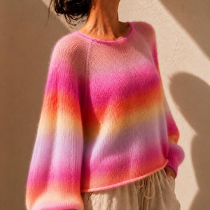 Könnte beinhalten: Ein langärmeliger Pullover mit einem lebendigen Farbverlauf, der von Pink über Orange und Gelb bis hin zu hellem Violett reicht. Das Kleidungsstück hat einen Rundhalsausschnitt, Raglanärmel und eine weiche, flauschige Textur im gesamten Strick.
