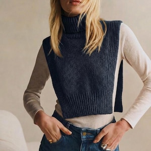 May include: A navy blue knitted turtleneck vest. The vest is worn over a long-sleeved beige shirt and paired with blue jeans. The model is wearing rings on their fingers.