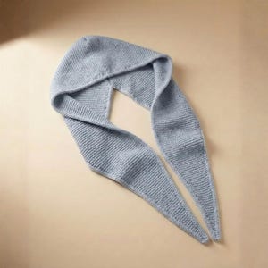 May include: A pale blue knitted hood and scarf. The hood is attached to a long scarf, forming a continuous piece. The knitted texture is clearly visible, and the scarf tapers to two pointed ends. The item is made of soft material.