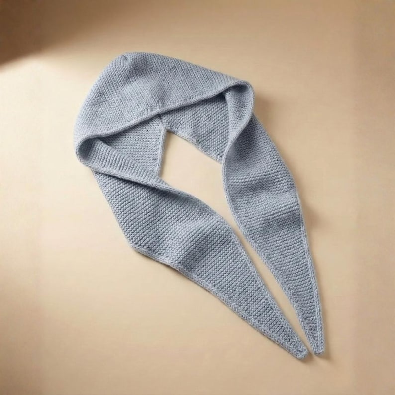 May include: A pale blue knitted hood and scarf. The hood is attached to a long scarf, forming a continuous piece. The knitted texture is clearly visible, and the scarf tapers to two pointed ends. The item is made of soft material.