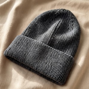 Puede incluir: Un gorro de punto gris oscuro con un borde doblado. El gorro está hecho de una tela de punto acanalado texturizado. El gorro está sobre una superficie de tela beige.