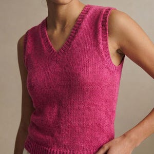Könnte beinhalten: Ein leuchtend fuchsiafarbener, ärmelloser Strickpullunder mit klassischem V-Ausschnitt und gerippten Abschlüssen an Kragen, Armausschnitten und Saum. Das Strickkleidungsstück hat eine texturierte, einfarbige Optik und wird über einer hellen Hose getragen.