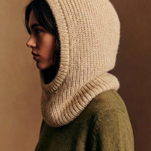 May include: A beige knit hood with a ribbed texture, worn over a green sweater. The hood frames the face, with a neck warmer that extends down to the shoulders. The neutral color palette creates a cozy and warm aesthetic.