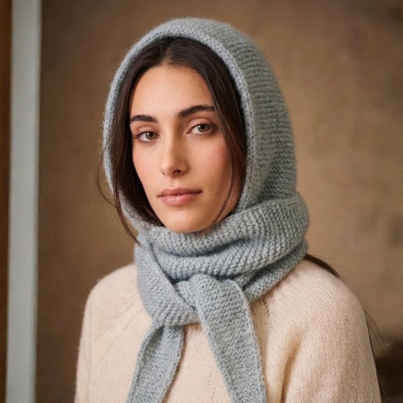 May include: A light blue knitted hood and scarf set. The hood frames the face, and the scarf is tied in a knot. The set is made of a soft, textured material. The model is wearing a cream-coloured jumper.