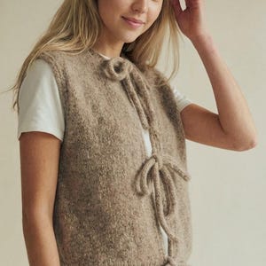 Könnte beinhalten: Beige Strickweste mit Bindebändern vorne. Die ärmellose Weste hat eine strukturierte Optik. Das Model trägt ein weißes T-Shirt und eine blaue Jeans. Die Weste ist neutral gefärbt und eignet sich zum Kombinieren.