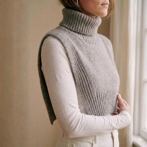 May include: A light gray knitted turtleneck vest with a ribbed texture. The vest is worn over a white long-sleeved shirt. The outfit is neutral-toned and appears to be made of soft, comfortable materials. The vest is a stylish layering piece.