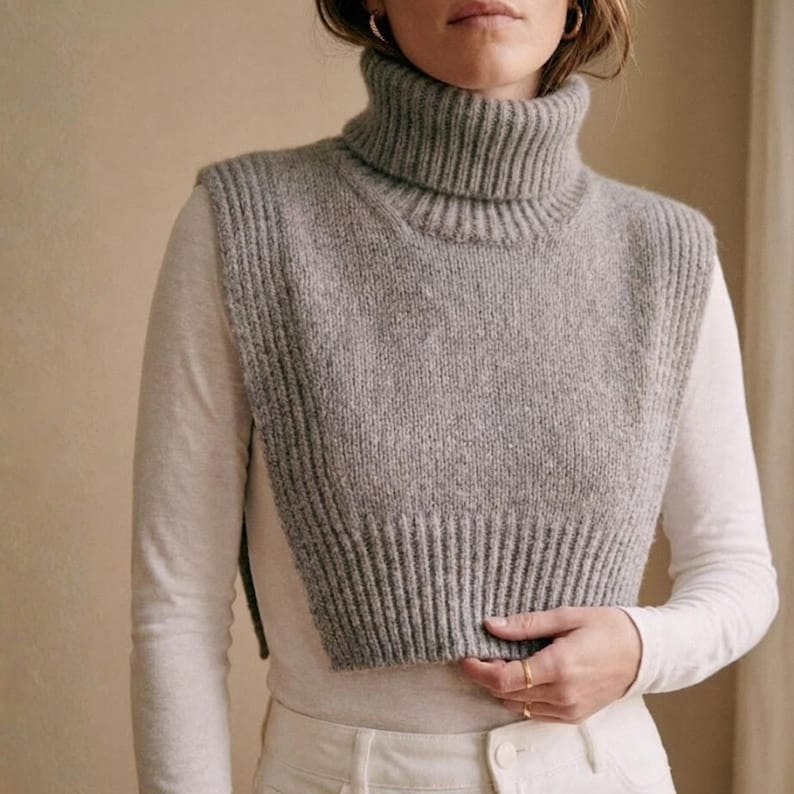May include: A gray knitted turtleneck collar and vest. The collar is ribbed and the vest has a textured knit design. The garment is worn over a white long-sleeved shirt and white pants. The overall style is minimalist and cozy.