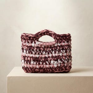 Könnte beinhalten: Quadratische Häkeltasche in Burgunderrot, Rosa und Weiß. Sie hat einen einzelnen, abgerundeten Griff. Das Häkelmuster erzeugt einen strukturierten, gewebten Effekt. Die Tasche steht auf einer neutralen Oberfläche. Ungefähre Abmessungen: 20 cm x 20 cm.