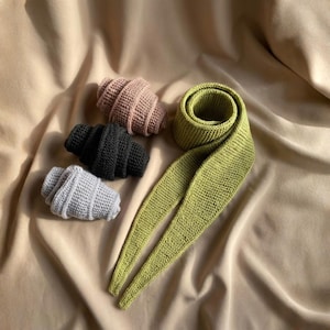Op de afbeelding: Een assortiment handgebreide items, waaronder drie croissantvormige objecten in roze, zwart en grijs, en een lange, groene gebreide sjaal. De items zijn gerangschikt op een beige stoffen achtergrond.