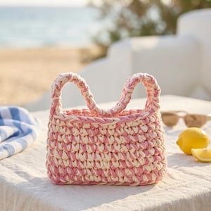 Könnte beinhalten: Kleine, handgehäkelte Tragetasche in Rosa und Creme. Die Tasche hat zwei stabile Griffe und eine rechteckige Form. Die Tasche steht auf einer weißen Oberfläche, mit einer Strandszene im Hintergrund.
