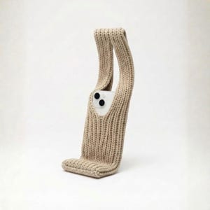 Könnte beinhalten: Eine beige gestrickte Handyhülle mit einem einzigartigen Design. Die Hülle hat eine rechteckige Basis und zwei lange, gebogene Griffe. Ein weißes Telefon ist im Inneren der Hülle sichtbar, mit seinen Kameralinsen. Die Hülle besteht aus dickem, strukturiertem Garn.