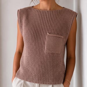Könnte beinhalten: Ein ärmelloses Strickoberteil in Mauve mit einer vertikalen Rippenstruktur am gesamten Körper. Das Kleidungsstück verfügt über eine einzelne aufgesetzte Tasche auf der linken Brustseite und einen weiten U-Boot-Ausschnitt mit einem geraden Saum.