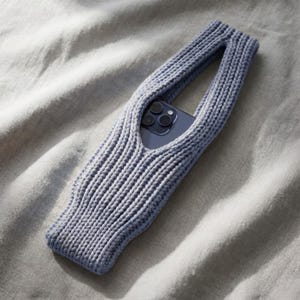 Könnte beinhalten: Eine hellblaue gestrickte Handytasche mit Griff, die ein Smartphone hält. Die Hülle besteht aus einem Rippenstrickstoff. Die Handytasche ist etwa 18 cm lang.
