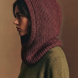 Puede incluir: Una capucha de punto burdeos con textura acanalada, usada sobre un suéter verde. La capucha enmarca la cara, creando un look cálido y acogedor. La paleta de colores es terrosa y otoñal.