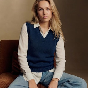 Könnte beinhalten: Eine marineblaue V-Ausschnitt-Weste über einem weißen Hemd mit Kragen und hellblauen Jeans. Die Weste ist einfarbig und scheint aus Strickmaterial zu sein. Das Model sitzt auf einem braunen Stuhl.