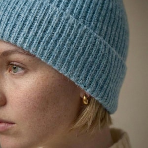 Pode incluir: Um close-up de um gorro de malha canelada azul claro. O gorro é feito de um material macio e texturizado. Um brinco dourado em forma de gota é visível. A pele da pessoa tem sardas e pele clara.
