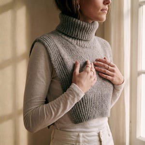 May include: A gray, ribbed knit turtleneck vest. The vest is worn over a long-sleeved, light gray shirt. The model is wearing rings on their fingers. The vest is a cropped style, with a high turtleneck collar.