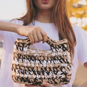Könnte beinhalten: Eine kleine, rechteckige Handtasche aus dickem, grobem Garn mit einem Streifenmuster in Dunkelbraun, Beige und Weiß. Die Tasche verfügt über einen stabilen, integrierten Griff an der Oberseite, der in derselben Häkelstruktur gefertigt ist.