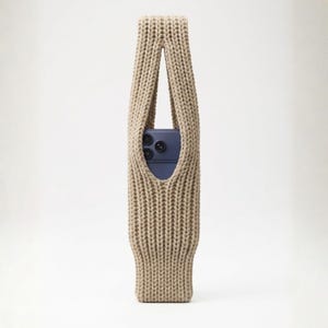 May include: A beige knitted phone sleeve with a handle. The sleeve is made of a thick, textured yarn and has a rectangular opening to display a blue phone. The phone has three camera lenses.
