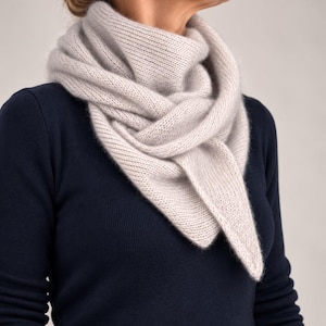 May include: A soft, light gray triangular knit scarf with a ribbed texture, draped around the neck. The scarf is worn over a dark navy blue long-sleeved sweater with a ribbed neckline and cuffs, creating a layered look with neutral tones.