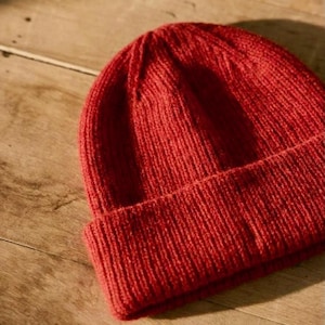 Puede incluir: Un gorro de punto rojo vibrante. El gorro está hecho de un material de punto acanalado suave. El gorro es de color liso y tiene un borde doblado. Es un accesorio de invierno.