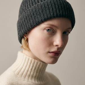 May include: A dark grey ribbed knit beanie hat. The hat is worn with a cream-coloured turtleneck sweater. The beanie is a classic style, suitable for cold weather. The hat is made of a soft, warm material.