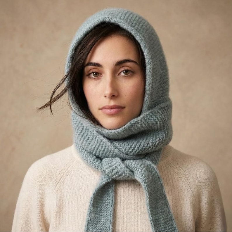 May include: A light blue knitted hood and scarf. The hood frames the face, and the scarf is knotted at the neck. The knitted texture is visible, and the colour is a soft, muted blue. The person is wearing a cream-coloured jumper.