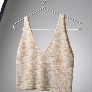 Könnte beinhalten: Ein ärmelloses Strick-Crop-Top mit tiefem V-Ausschnitt und breiten Trägern. Das Kleidungsstück besteht aus einem melierten Garnmix in Creme-, Beige- und hellen Brauntönen, was dem Stoff ein strukturiertes und gesprenkeltes Aussehen verleiht.