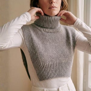 May include: A gray knitted turtleneck collar, worn over a white long-sleeved shirt. The turtleneck is a neutral gray color and appears to be made of a soft, textured yarn. The collar is a versatile accessory.