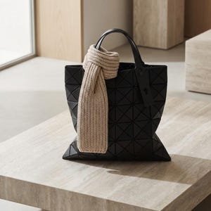Puede incluir: Un bolso tote negro geométrico con un asa a juego y una bufanda beige sobre él. La bolsa está sobre una superficie de color claro. La bufanda es de color beige claro.