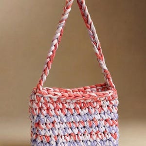 May include: A handmade tote bag in a woven design. The bag features a rectangular body with a braided handle. The color scheme includes white, coral, and lavender hues, creating a soft, textured appearance. The bag is suitable for carrying various items.