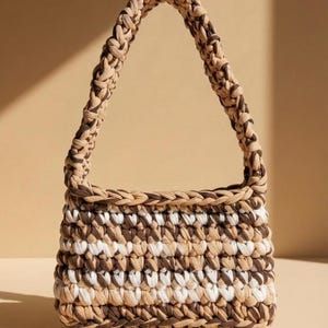 Könnte beinhalten: Eine handgefertigte Häkeltasche in Beige-, Braun- und Weißtönen. Die Tasche hat einen rechteckigen Körper und einen geflochtenen Griff. Das Häkelmuster erzeugt einen strukturierten, gestreiften Effekt. Die Tasche ist für den Transport von Essentials konzipiert.