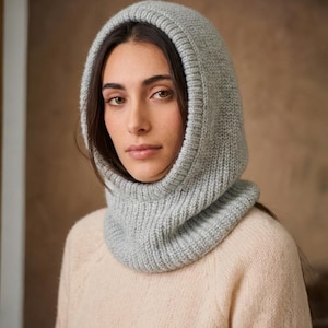 Könnte beinhalten: Eine hellgraue Strickhaube, die Kopf und Hals bedeckt. Die Kapuze ist aus weichem, strukturiertem Garn gefertigt und hat ein Rippendesign um Gesicht und Hals. Das Model trägt einen hellpfirsichfarbenen Pullover.