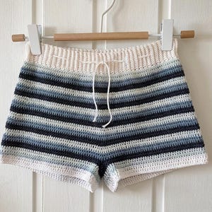 May include: Hand-crocheted shorts featuring horizontal stripes in blue, grey, and white. The shorts have a drawstring waist and are displayed on a wooden hanger. The crochet work is detailed and the colour palette is neutral.