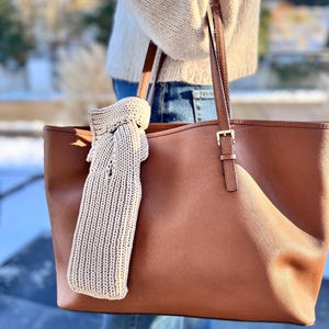 Könnte beinhalten: Eine große, braune Ledertasche mit passendem Riemen und goldfarbener Schnalle. Ein beiger Strickschal hängt an der Tasche. Die Tasche wird von einer Person getragen, die einen beigen Pullover und eine blaue Jeans trägt.