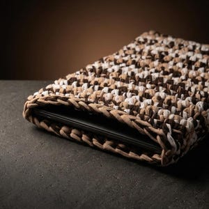 Könnte beinhalten: Eine rechteckige Laptoptasche aus dickem, geflochtenem Stoff in einem melierten Muster aus Dunkelbraun, Beige und Weiß. Die Tasche weist ein grobes, strukturiertes Häkelmuster auf und enthält ein flaches, schwarzes elektronisches Gerät.