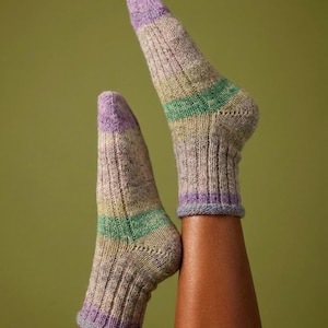 Könnte beinhalten: Ein Paar gestrickte Socken mit Rippenmuster. Die Socken haben ein Farbblockmuster mit lila Zehen und Bündchen, einen grünen Streifen und eine neutrale Grundfarbe. Die Socken werden vor einem grünen Hintergrund präsentiert.