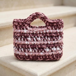 Könnte beinhalten: Eine quadratische, handgefertigte Häkeltasche in den Farbtönen Burgunder, Rosa und Weiß. Die Tasche hat einen stabilen Griff und ein strukturiertes, gewebtes Aussehen. Die Tasche steht auf einer Steinoberfläche.