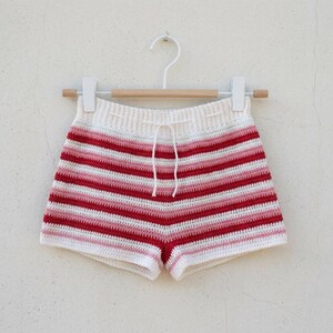 Könnte beinhalten: Häkelshorts mit horizontalen Streifen in Weiß, Hellrosa und Dunkelrot. Die Shorts haben ein hoch tailliertes Design mit geripptem Bund und einer weißen Kordel und hängen an einem Holzkleiderbügel mit weißen Clips.
