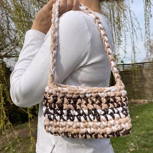 Könnte beinhalten: Eine rechteckige Handtasche mit geflochtenem Riemen. Die Tasche besteht aus einem gewebten Stoff in Braun-, Beige- und Weißtönen. Der Riemen ist ein geflochtenes Design in Weiß und Beige.