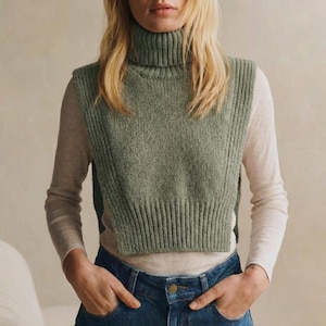 May include: A sage green turtleneck sweater vest with ribbed detailing. The sleeveless vest is paired with a long-sleeved, light-colored shirt and blue jeans. The vest has a high, folded collar.