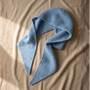 Könnte beinhalten: Eine hellblaue Strickhaube mit langen, spitz zulaufenden Schalenden. Die Kapuze und der Schal sind aus einem strukturierten Strickstoff gefertigt. Die Schalenden haben einen dekorativen Rand. Der Artikel wird auf einer neutralfarbenen Oberfläche präsentiert.