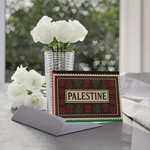 Puede incluir: Una tarjeta de felicitación rectangular con un fondo oscuro, un patrón floral geométrico en rojo y verde y la palabra PALESTINE en un recuadro color crema en el centro. La tarjeta está junto a un florero de cristal con rosas blancas.