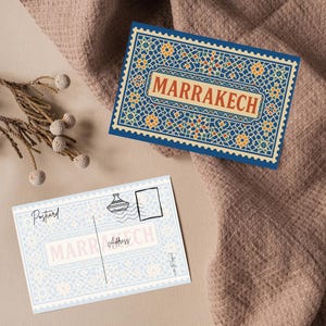 May include: Two postcards with a Moroccan theme. One postcard features the word "MARRAKECH" in a rectangular frame with a blue, orange, and white geometric pattern. The other postcard has the word "MARRAKECH" and the word "Postcard" printed on it.