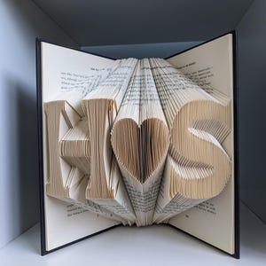 May include: A book sculpture with the word "HIS" and a heart shape carved into the pages. The book is open, with the letters and heart standing out. The pages are beige, and the book has a black cover.